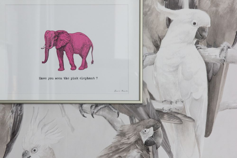 Auf einer Wand mit Papageientapete in Sepiatönen hängt ein gerahmtes Bild mit einem rosa Elefanten.