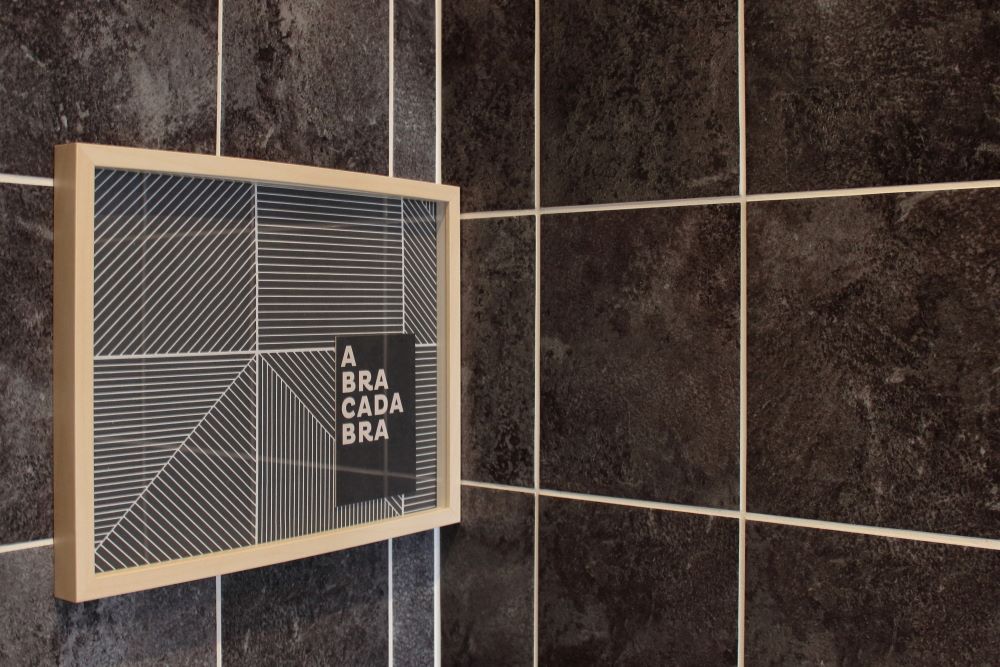 Ein Bilderrahmen aus hellem Birkenholz auf dunkler Wand sorgt für maximale Gemütlichkeit im Badezimmer. Gerahmt ist ein Bild namens Abracadabra, als Passpartout dient die Tapete des Raumes.