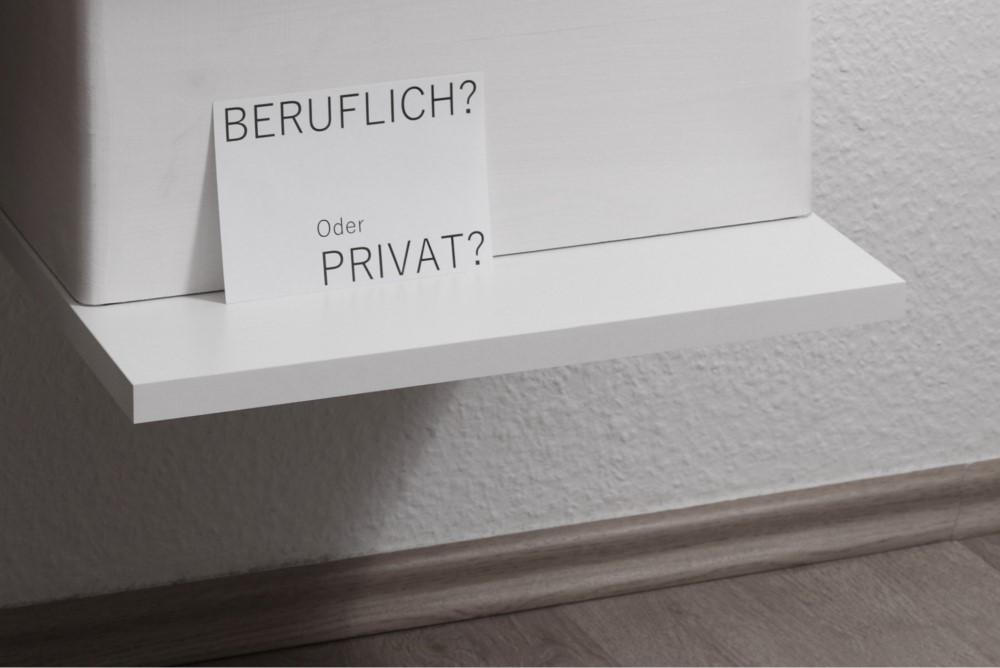 Ein weißes Regal mit einer weißen Holzkiste, an der eine Postkarte mit der Aufschrift "BERUFLICH oder PRIVAT" lehnt.