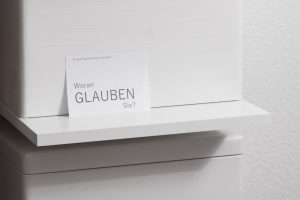 Ein weißes Regal mit einer weißen Holzkiste, an der eine Postkarte mit der Aufschrift "Woran GLAUBEN Sie" lehnt.