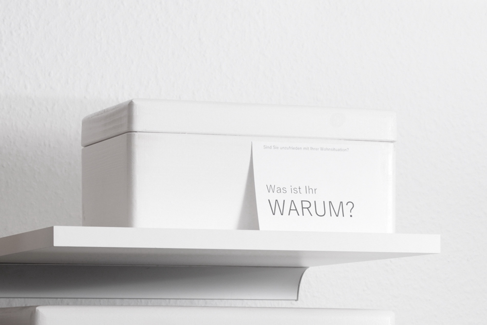 Ein weißes Regal mit einer weißen Holzkiste, an der eine Postkarte mit der Aufschrift "Was ist Ihr WARUM" lehnt.