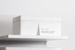 Ein weißes Regal mit einer weißen Holzkiste, an der eine Postkarte mit der Aufschrift "Was ist Ihr WARUM" lehnt.