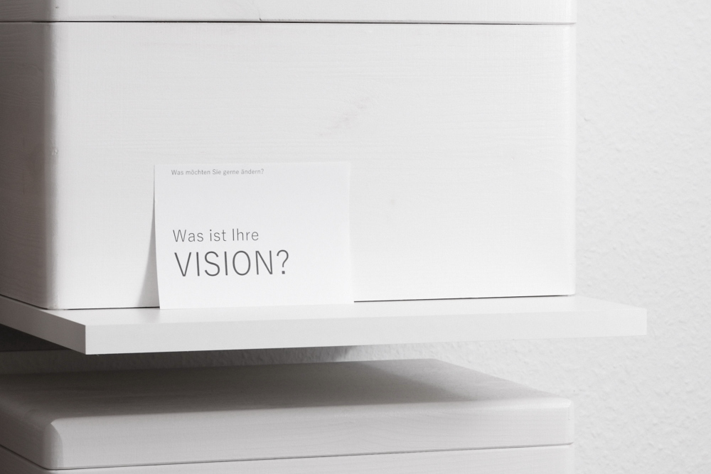 Ein weißes Regal mit einer weißen Holzkiste, an der eine Postkarte mit der Aufschrift "Was ist Ihre VISION" lehnt.