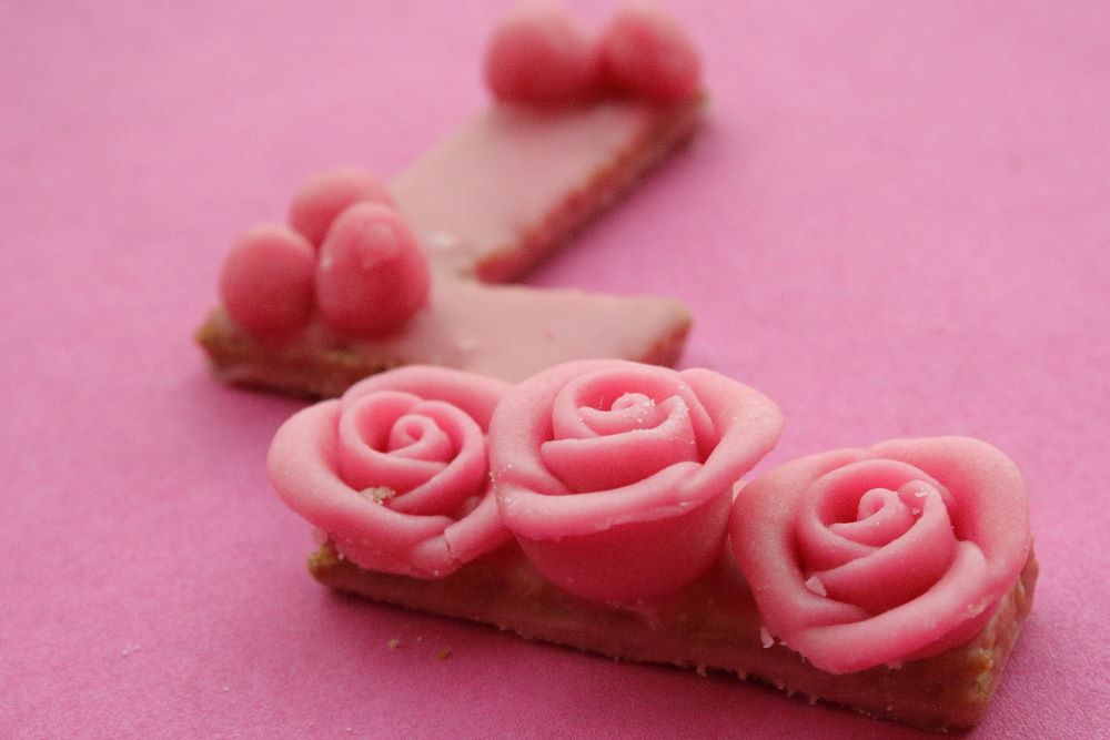 Buchstaben-Plätzchen mit rosa Zuckerguss und rosa Rosenblüten aus Marzipan.
