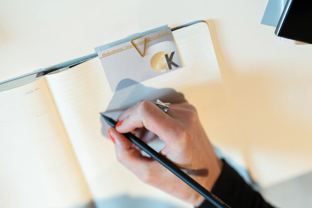 Eine Frauenhand notiert mit Bleistift etwas in einen Kalender, am Rand ist eine Visitenkarte der Firma diakubana innenraumgestaltung mit einer goldenen Büroklammer angeheftet. 